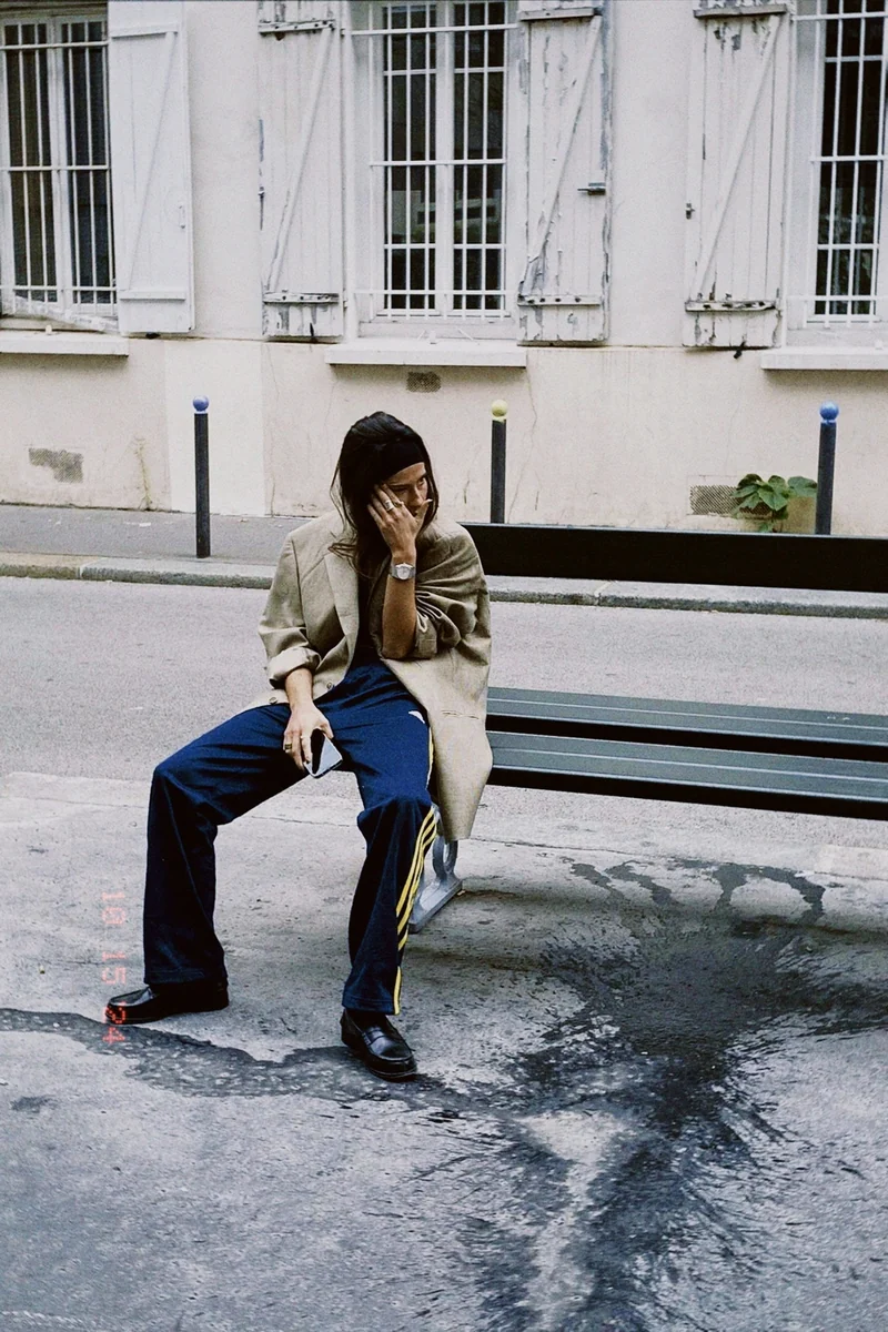 Valentine seated on a Paris street, wearing a beige blazer and navy trousers